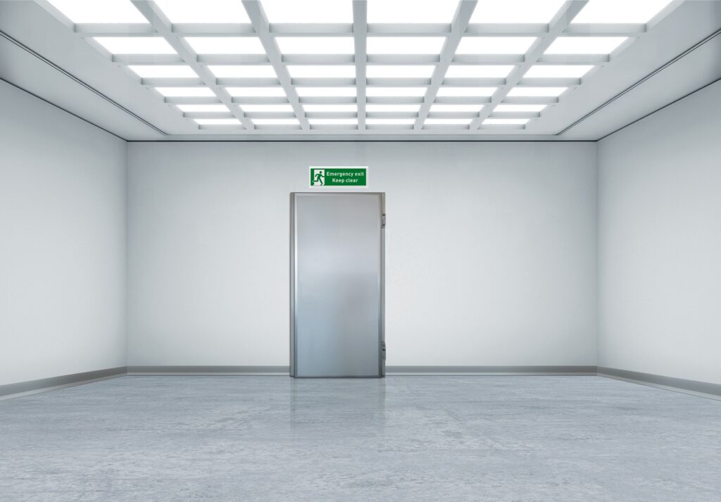 Empty white room with a central metal door and emergency exit sign.