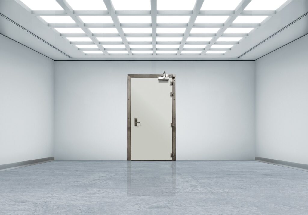 Clean, empty white room with a heavy steel door and bright grid ceiling lights.